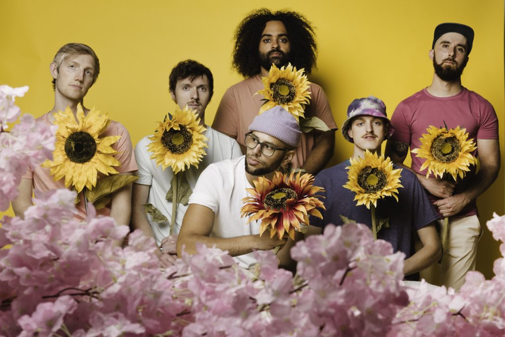 Joe Hertler and The Rainbow Seekers holding sunflowers in front of a yellow background.