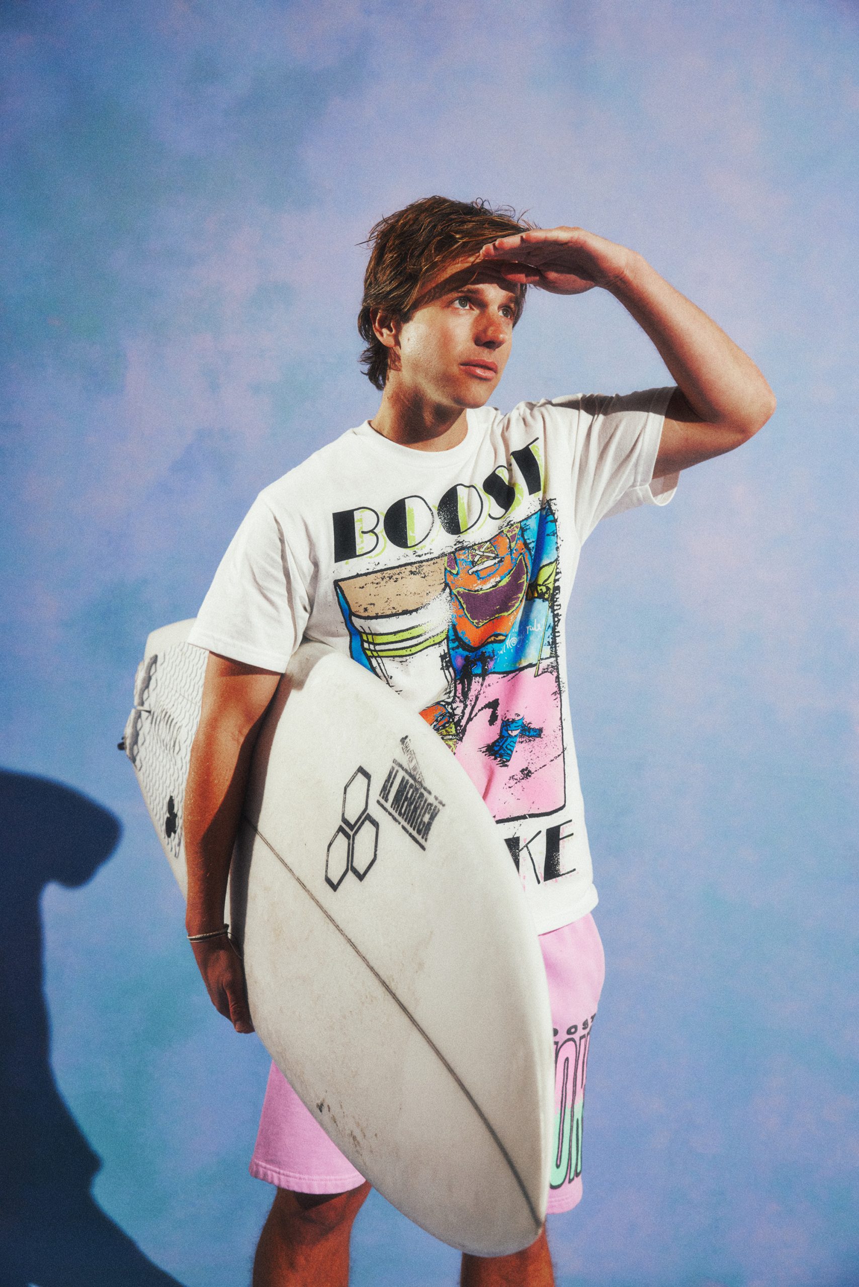 Chad Kroeger holding a surfboard in a retro Boost graphic tee against a blue sky backdrop