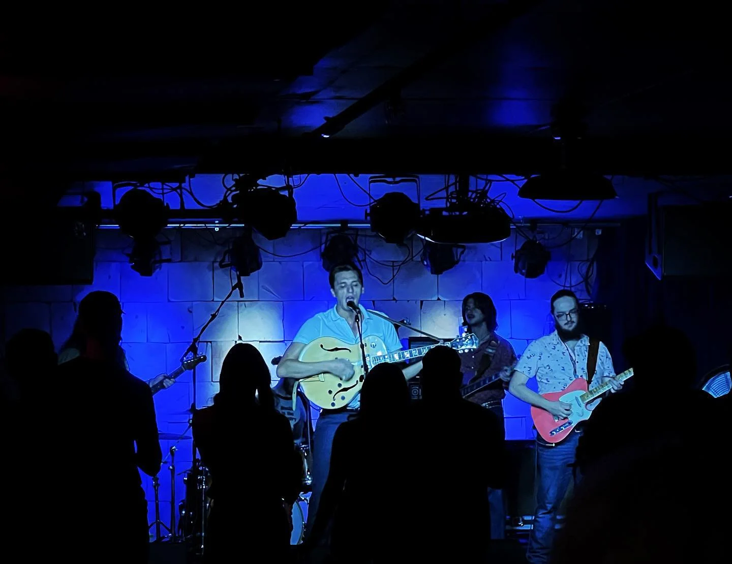 A band in western shirts plays under blue stage lights at The Black Buzzard as the crowd watches in silhouette.