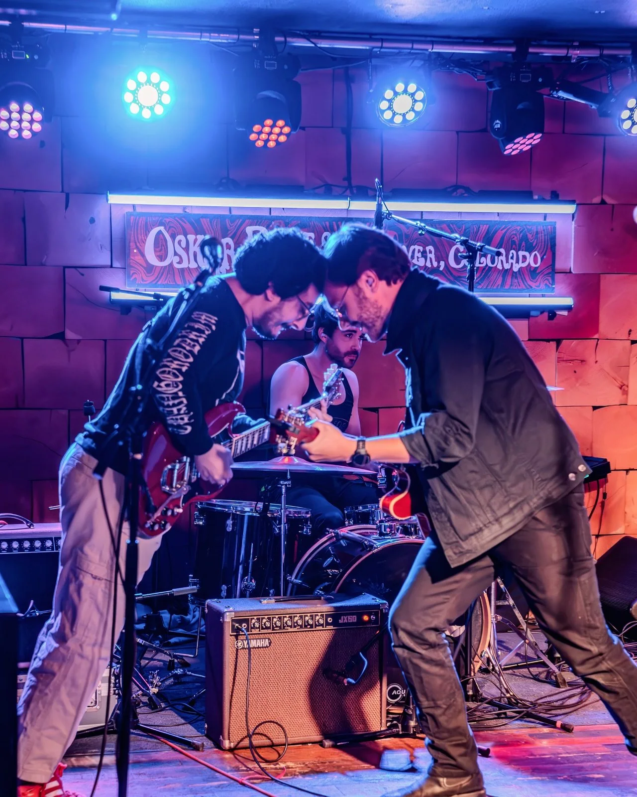 Two Hollow Head guitarists face off in front of the drummer under blue and red light.