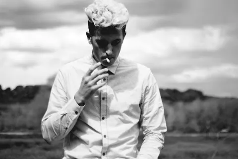 Black and white portrait of a man with bleached curly hair and a white shirt, smoking in a grassy field.