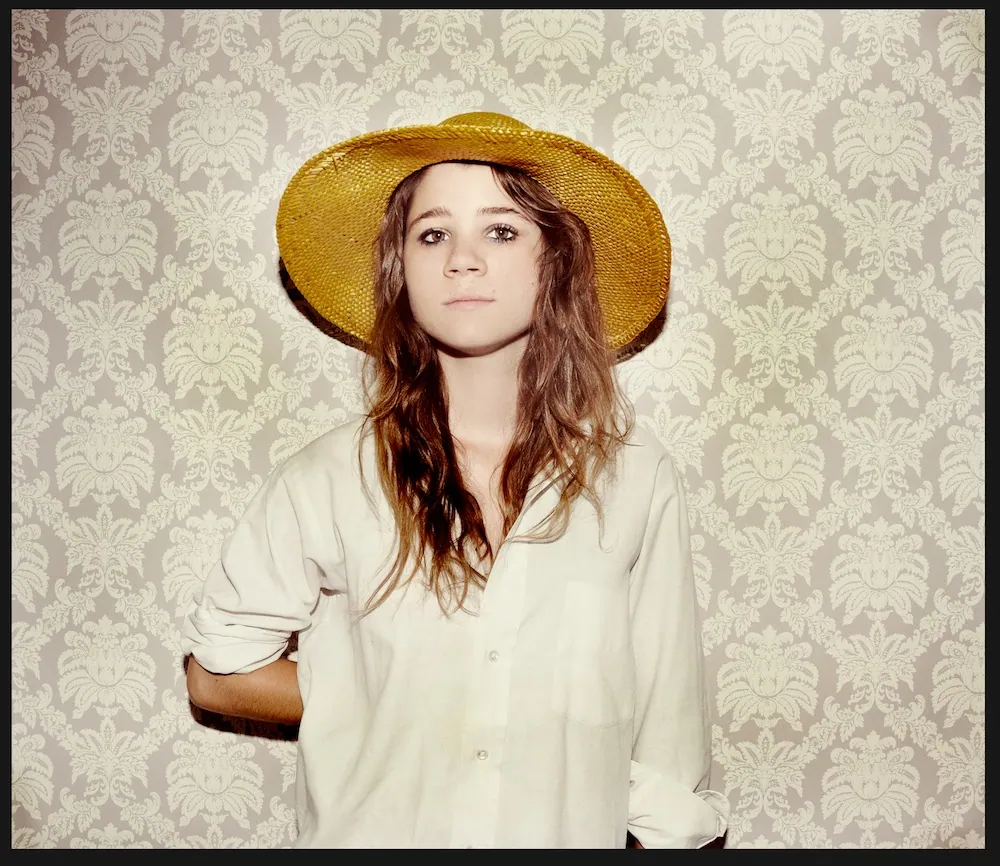 A woman with long brown hair wears a yellow straw hat and white button-up shirt against a cream damask wallpaper.