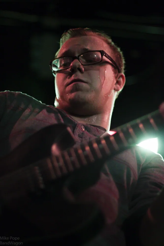 A sweaty guitarist in glasses plays an electric guitar under red stage lights during a battle of the bands.