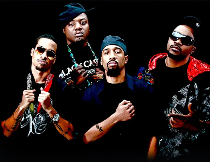 Four men pose against a black backdrop wearing hats, sunglasses, and chains, with fists raised.