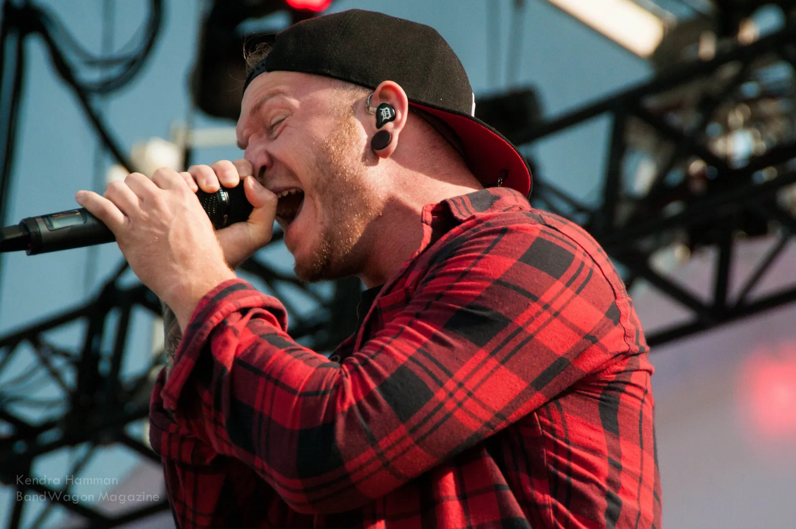 A singer in a backwards cap and red plaid shirt belts into a mic on a festival stage with metal truss behind.