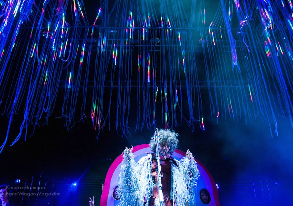 Performer with curly hair and silver fringe sings at a mic under hanging blue LED strands on stage.