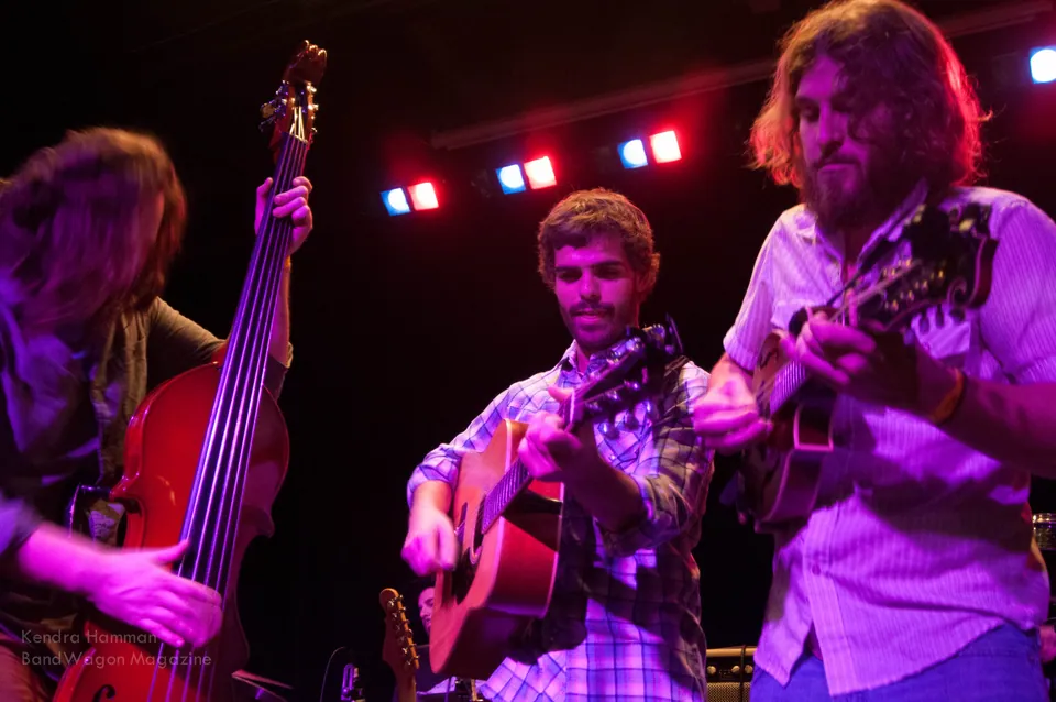 Three musicians perform on a dark stage under red and purple lights, with an upright bass and two acoustic guitars.
