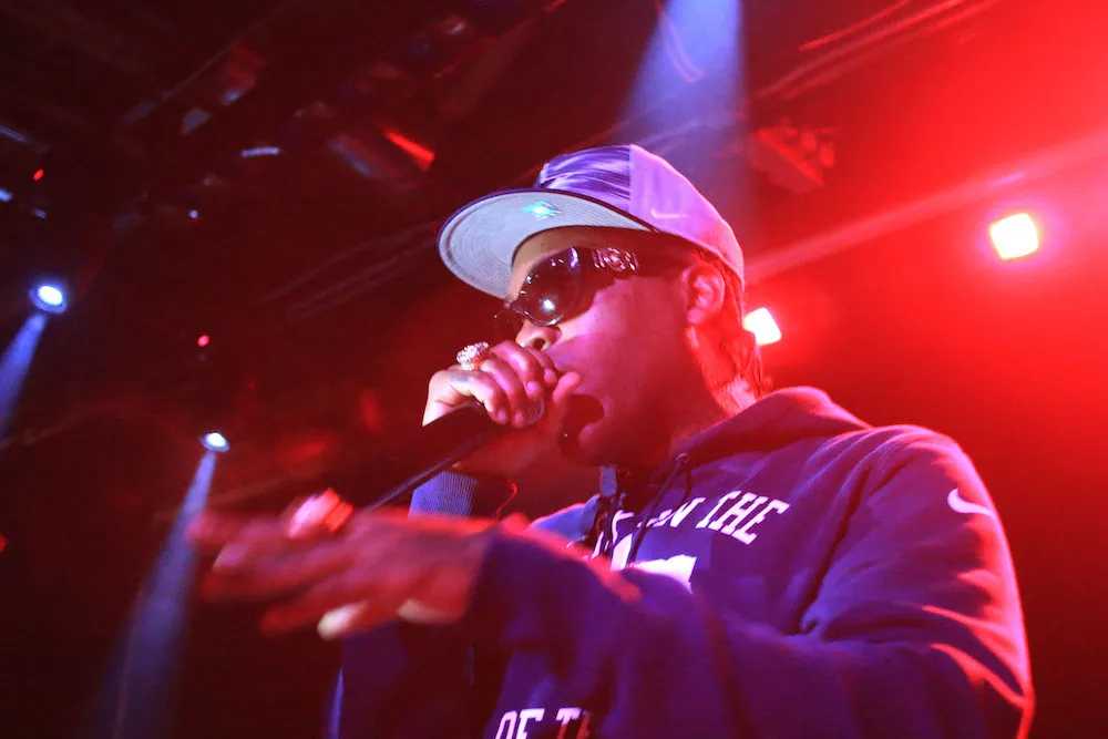 Mike Jones performs into a microphone under red stage lights, wearing sunglasses, a cap, and a purple hoodie.