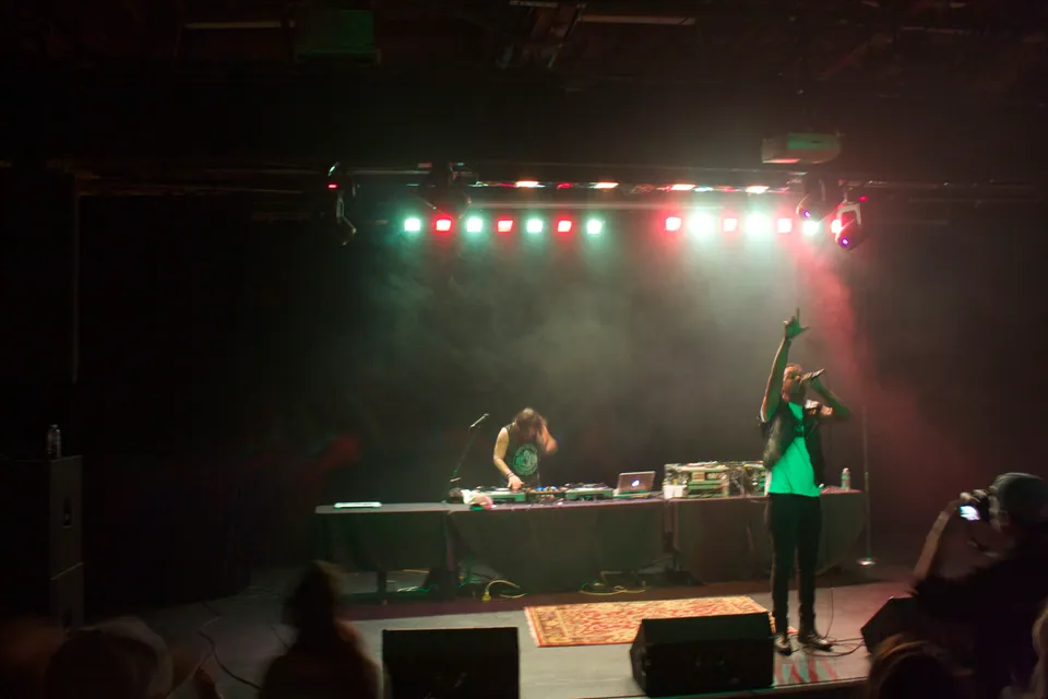 Pigeon John performs at the Moxi Theater as a DJ works turntables under red and green stage lights.