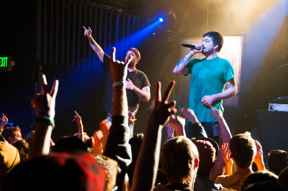 Two performers at the Aggie Theater, one in a teal shirt singing into a mic, as the crowd raises hands under blue stage