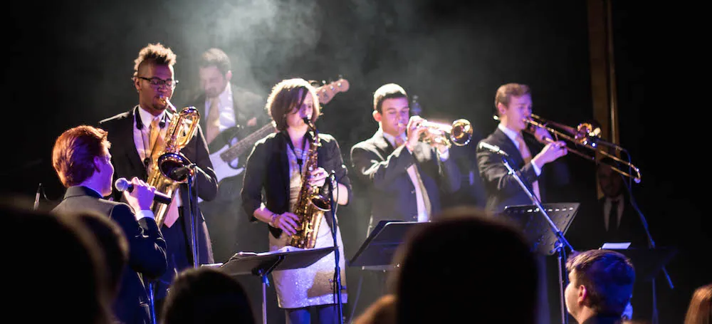 The Burroughs play on stage with saxophone, trumpet, trombone, and guitar as audience silhouettes watch.