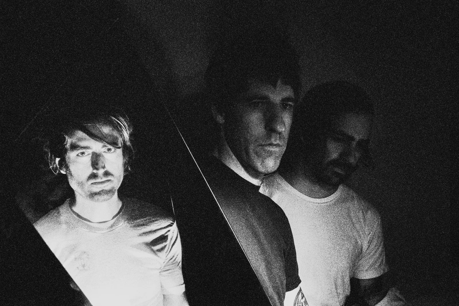Black and white portrait of three men in T-shirts against a dark background, one lit by harsh side light.