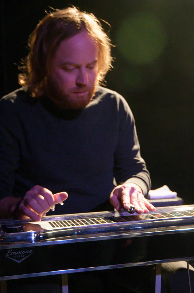 Bearded, red-haired musician in a dark sweater playing an Emmons pedal steel guitar on a dimly lit stage.