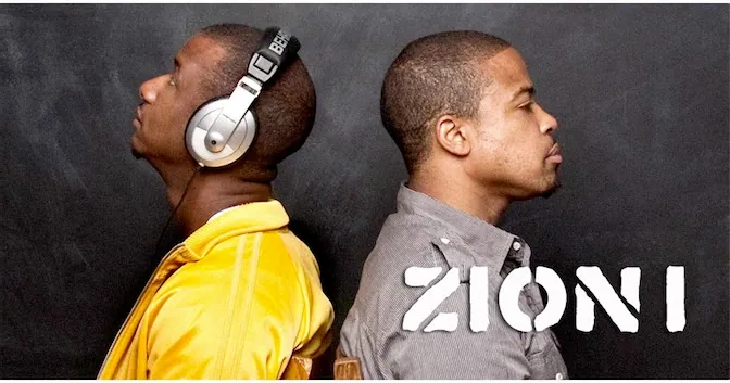 Zion I members pose back to back in profile against a dark chalkboard, one in a yellow jacket and headphones.