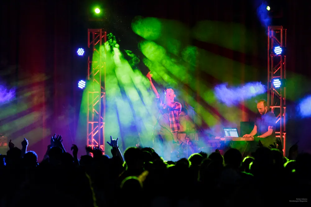 Crowd with hands raised faces a green-lit stage where a plaid-shirted performer stands beside a man at a laptop.