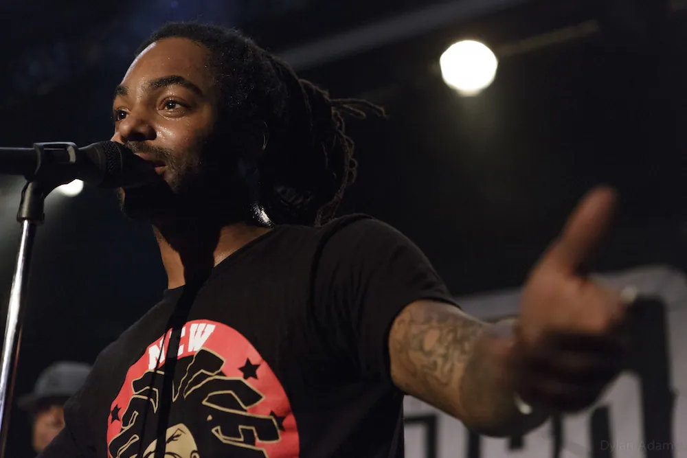 A dreadlocked performer sings into a microphone and points toward the crowd on a dimly lit stage at The Moxi.