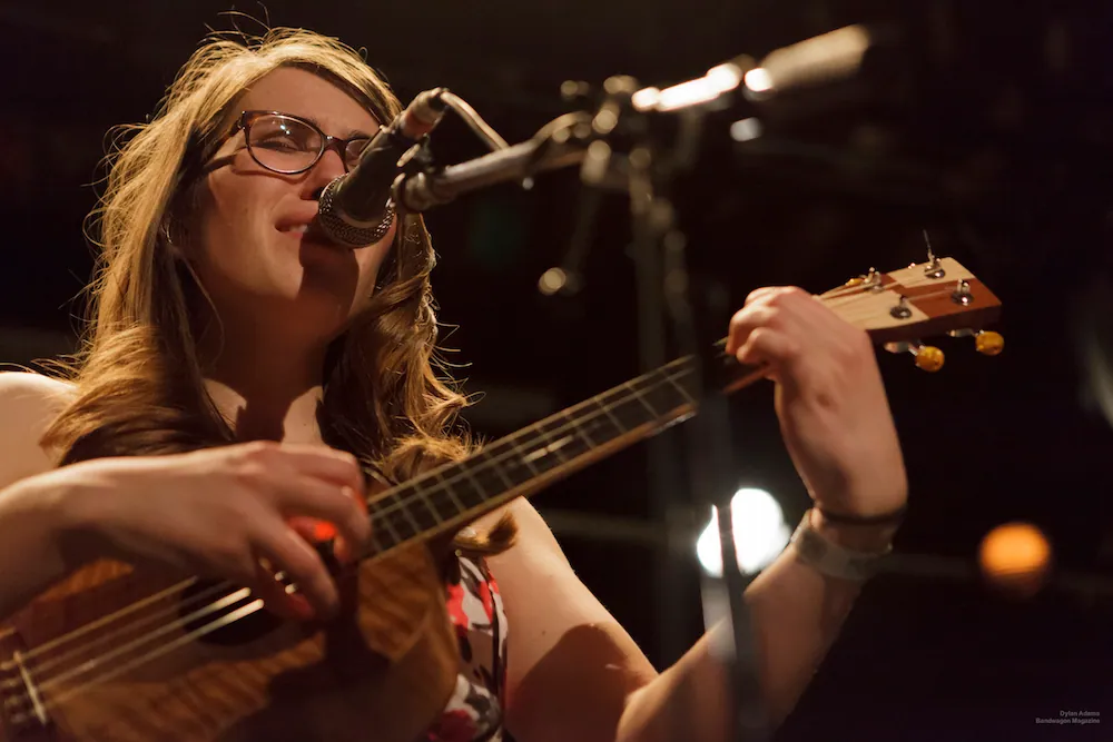 Woman with glasses and wavy hair sings into a microphone while playing a ukulele on a dimly lit stage.