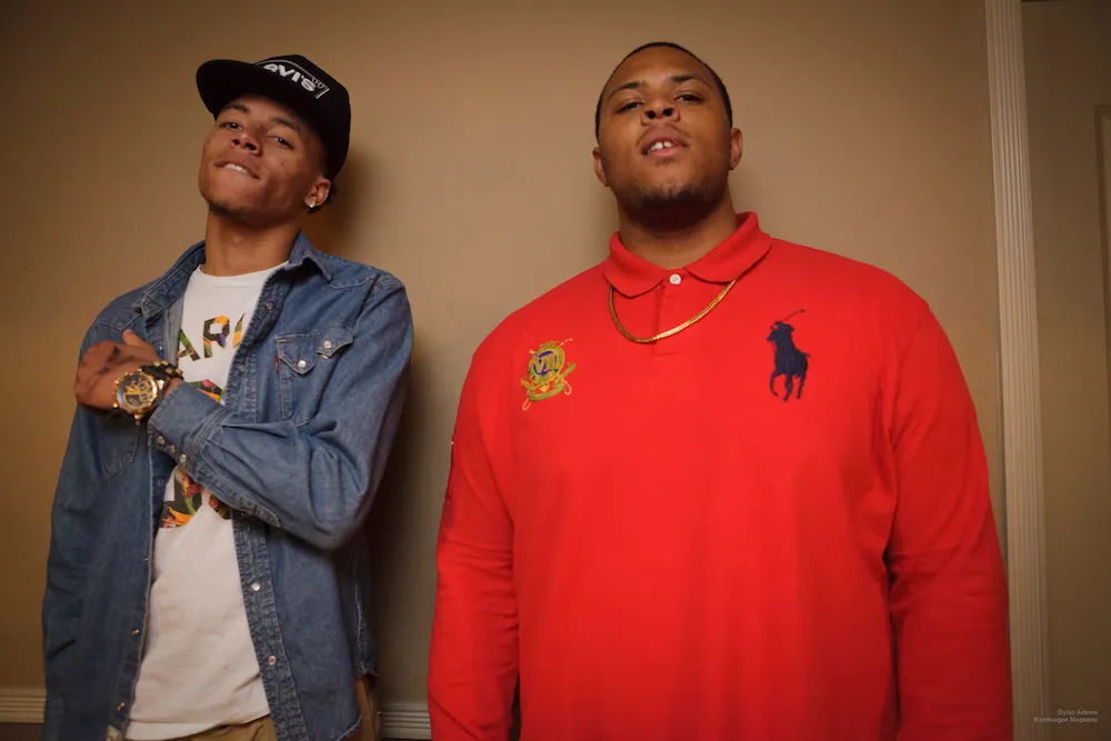 Two young men pose against a beige wall, one in a denim shirt and cap, the other in a red polo and gold chain.