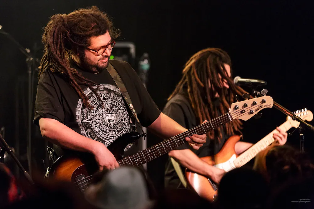 Two dreadlocked musicians play bass and electric guitar on a dark stage in front of a silhouetted crowd.