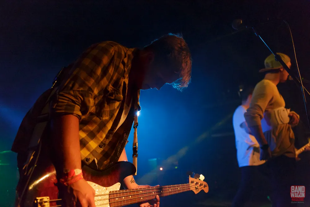 Bassist in plaid flannel leans over a bass guitar under blue stage lights as a cap-wearing musician performs behind.
