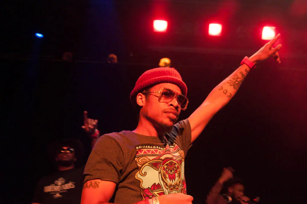 Performer in a red beanie and sunglasses raises an arm under red stage lights, with a crowd behind him.