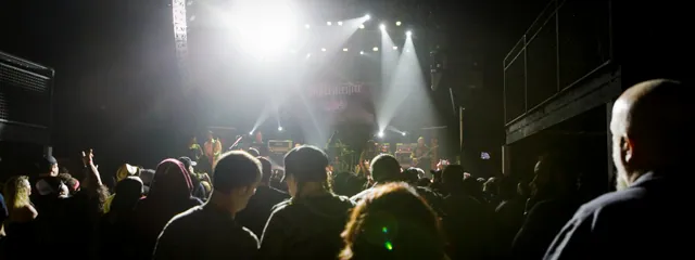 Concertgoers in silhouette face a bright stage with spotlights and beams cutting through haze at a dim music venue.