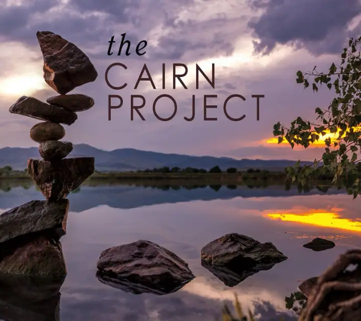 Balanced stone cairn by a calm lake at sunset, with mountains behind and "the CAIRN PROJECT" text overlaid.