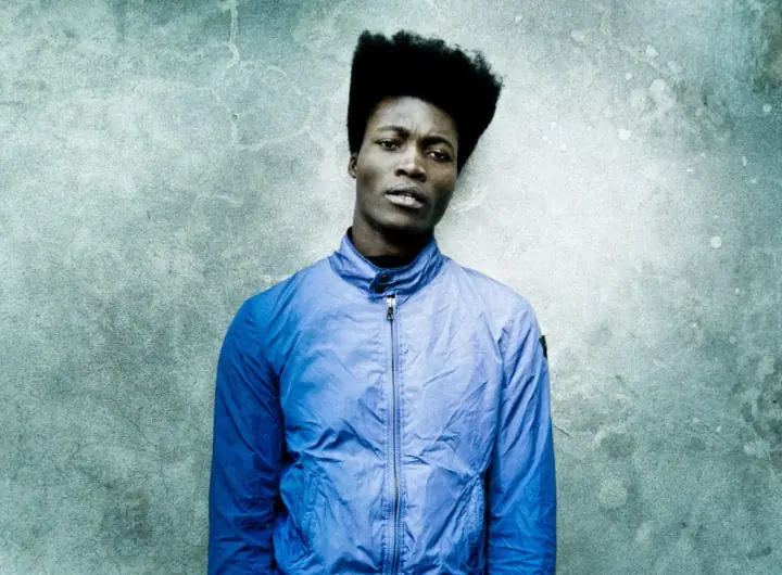 A man with voluminous natural hair wears a blue zip-up jacket against a weathered gray concrete wall.