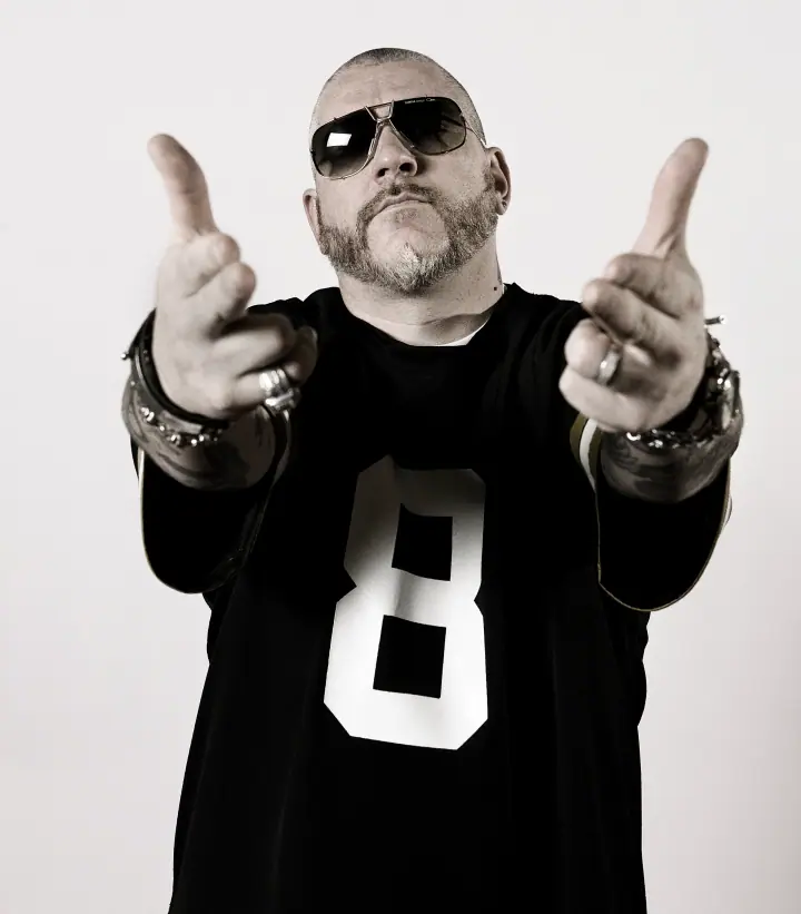 Man in aviator sunglasses and a black jersey with a large white 8 points both index fingers at the camera against a plain