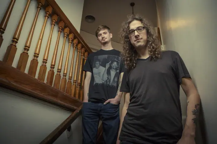 Two men stand on a wooden staircase, one in a graphic tee, the other with curly hair and glasses.