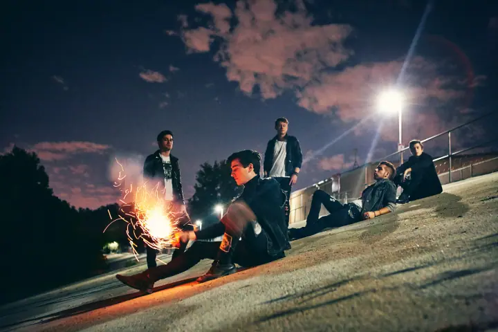 Five young men in dark jackets sit and stand on a concrete rooftop at night, one holding a lit sparkler.