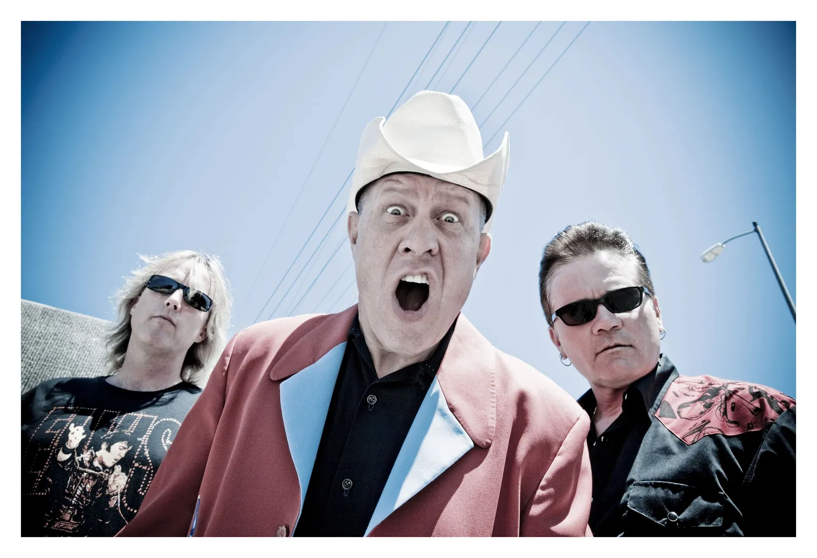 Three men pose outdoors under a blue sky; the center man in a white cowboy hat and pink jacket shouts, wide-eyed.