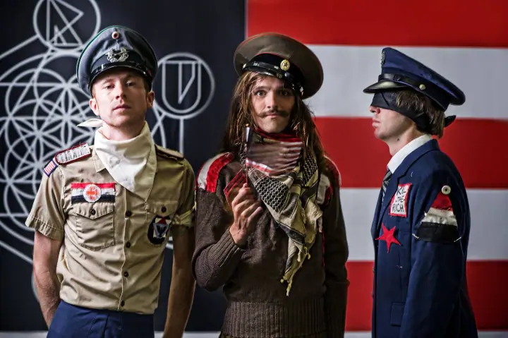 Three men in quirky military-style costumes stand before a red-and-white striped flag.