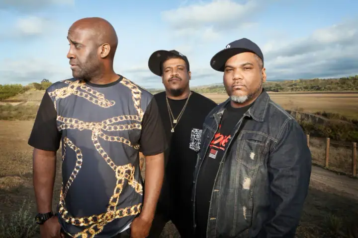 Three men in caps and casual streetwear pose outdoors in a dry grassy field under a partly cloudy sky.