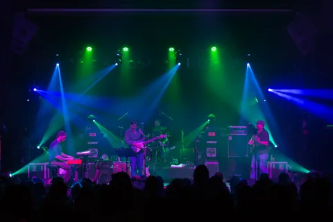 Dead Floyd performs under green and blue stage lights, with keyboards, guitar, and drums before a crowd.