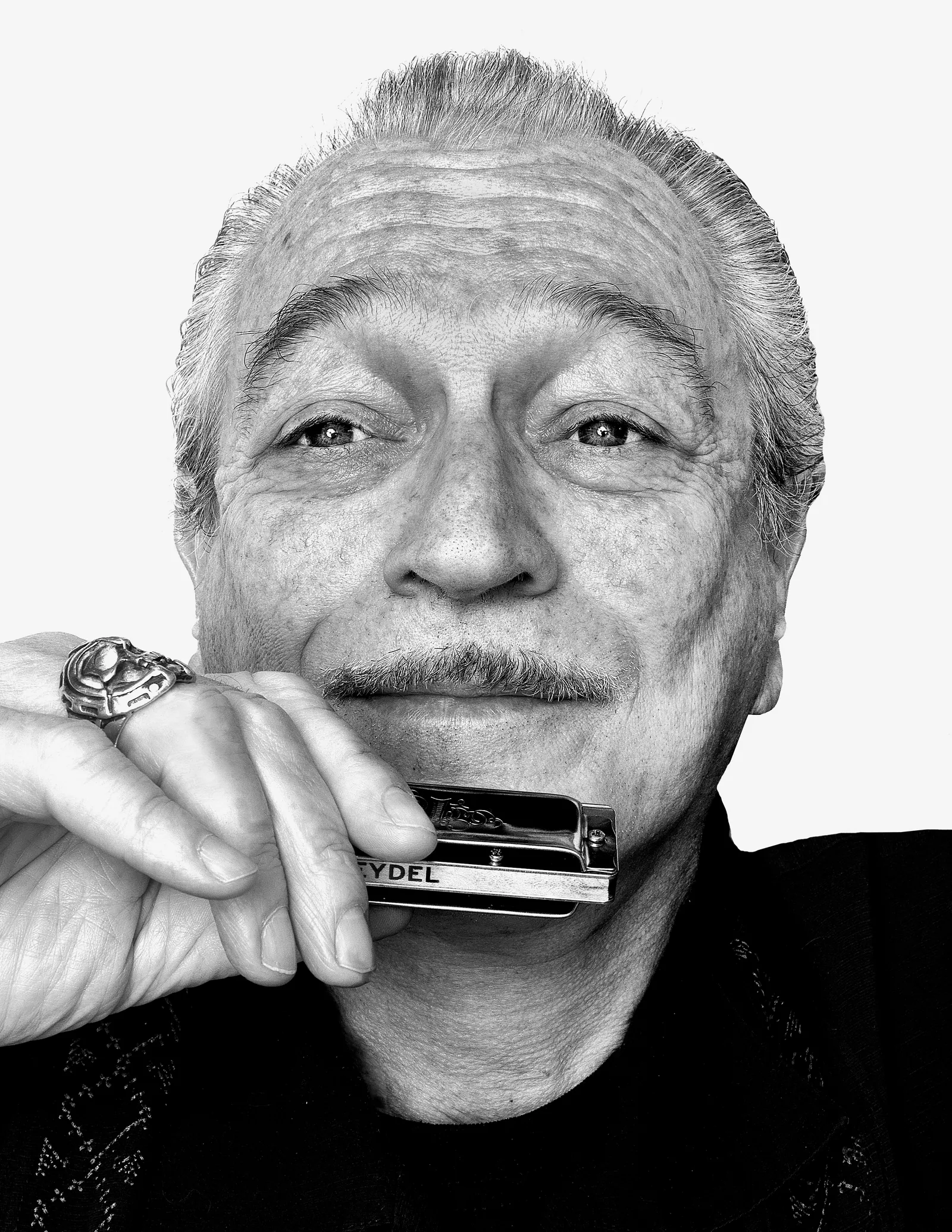 Black-and-white portrait of an older man with gray hair and a ring, playing a harmonica against a white background.