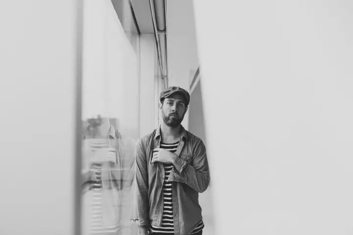 Black-and-white portrait of a bearded man in a flat cap and denim shirt over a striped top by a window.