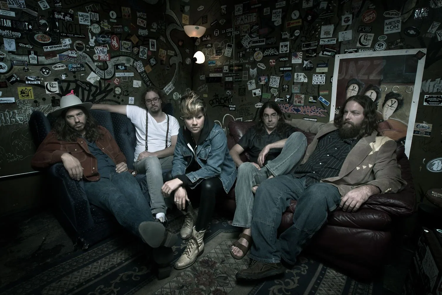 Five people pose on worn couches in a dim room covered with stickers, graffiti, and posters.
