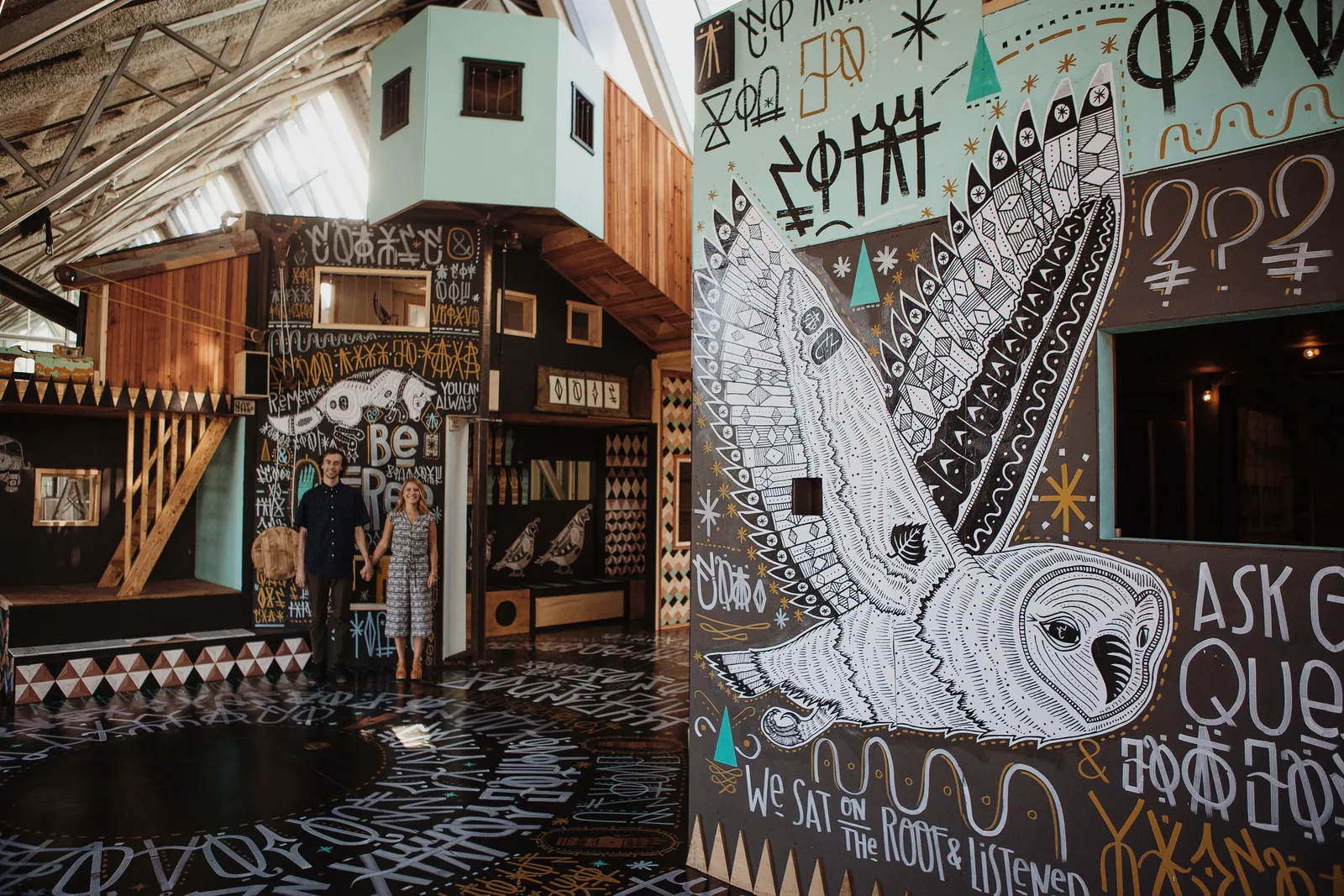 A couple stands hand in hand in a room covered with black-and-white hand-painted murals of an owl and symbols.