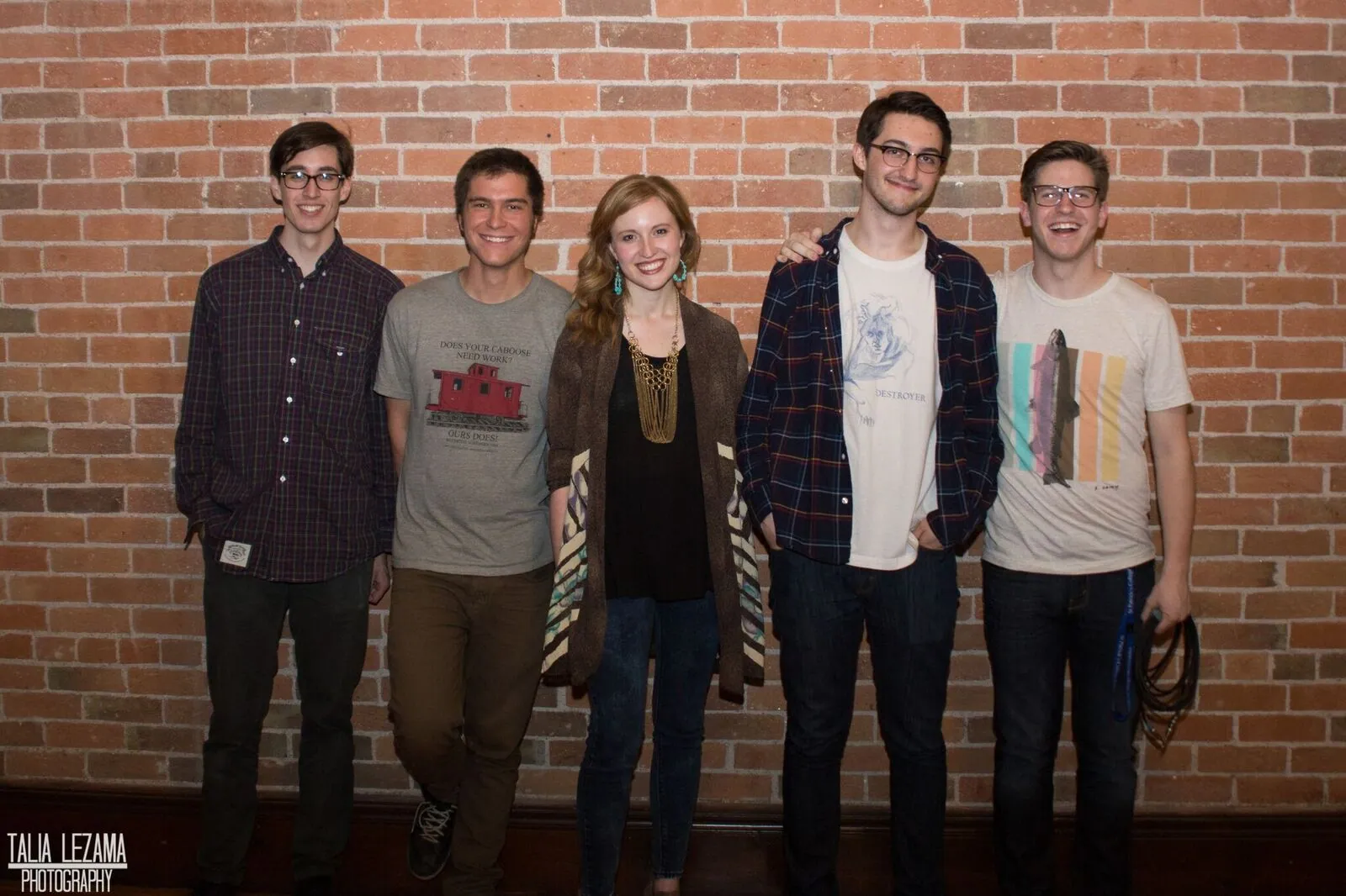 Five smiling people stand side by side against an exposed brick wall, with no instruments visible.