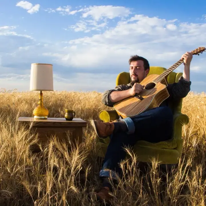Bearded man strums an acoustic guitar in a green armchair in a wheat field beside a lamp and mug.