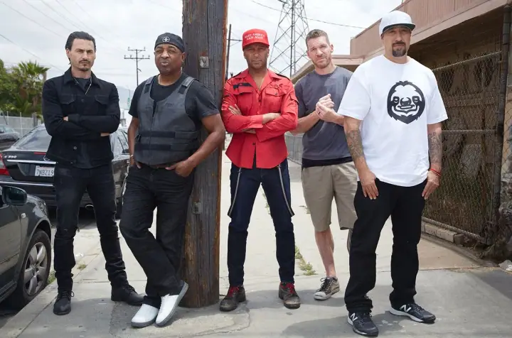 Five men pose on a city sidewalk beside a wooden utility pole, with parked cars and a chain-link fence behind them.