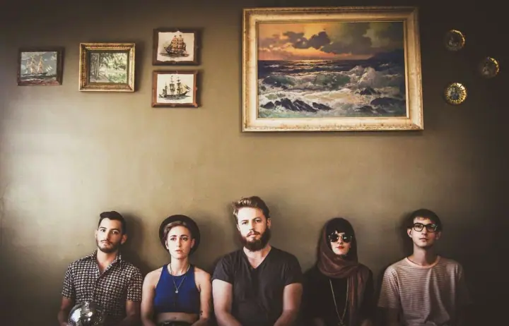 Five people sit in a row against a wall beneath a framed seascape painting and small ship pictures.