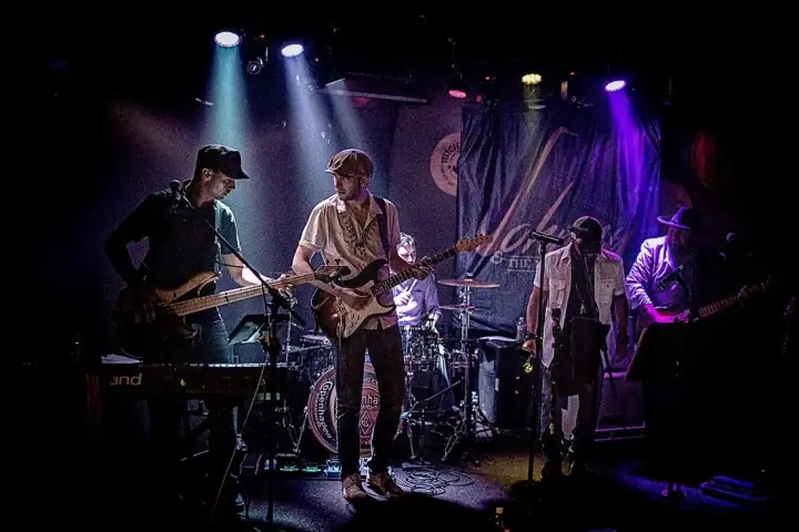 Five musicians perform on a dimly lit stage under blue and purple lights, with bass, guitar, keyboard, and drums.