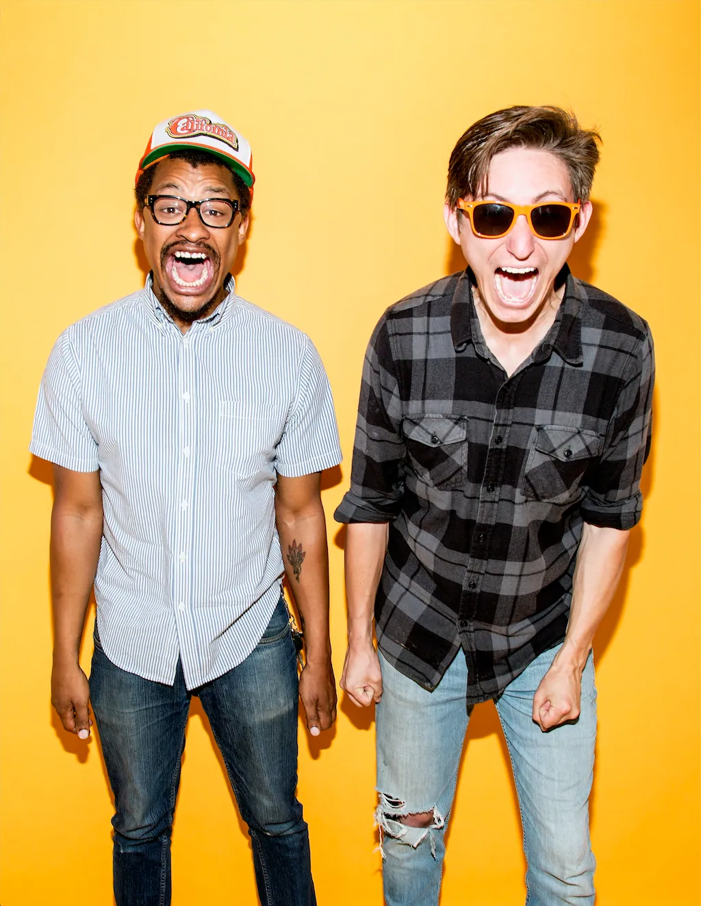 Two men shout excitedly against a yellow backdrop, one in a striped shirt and cap, one in orange sunglasses.