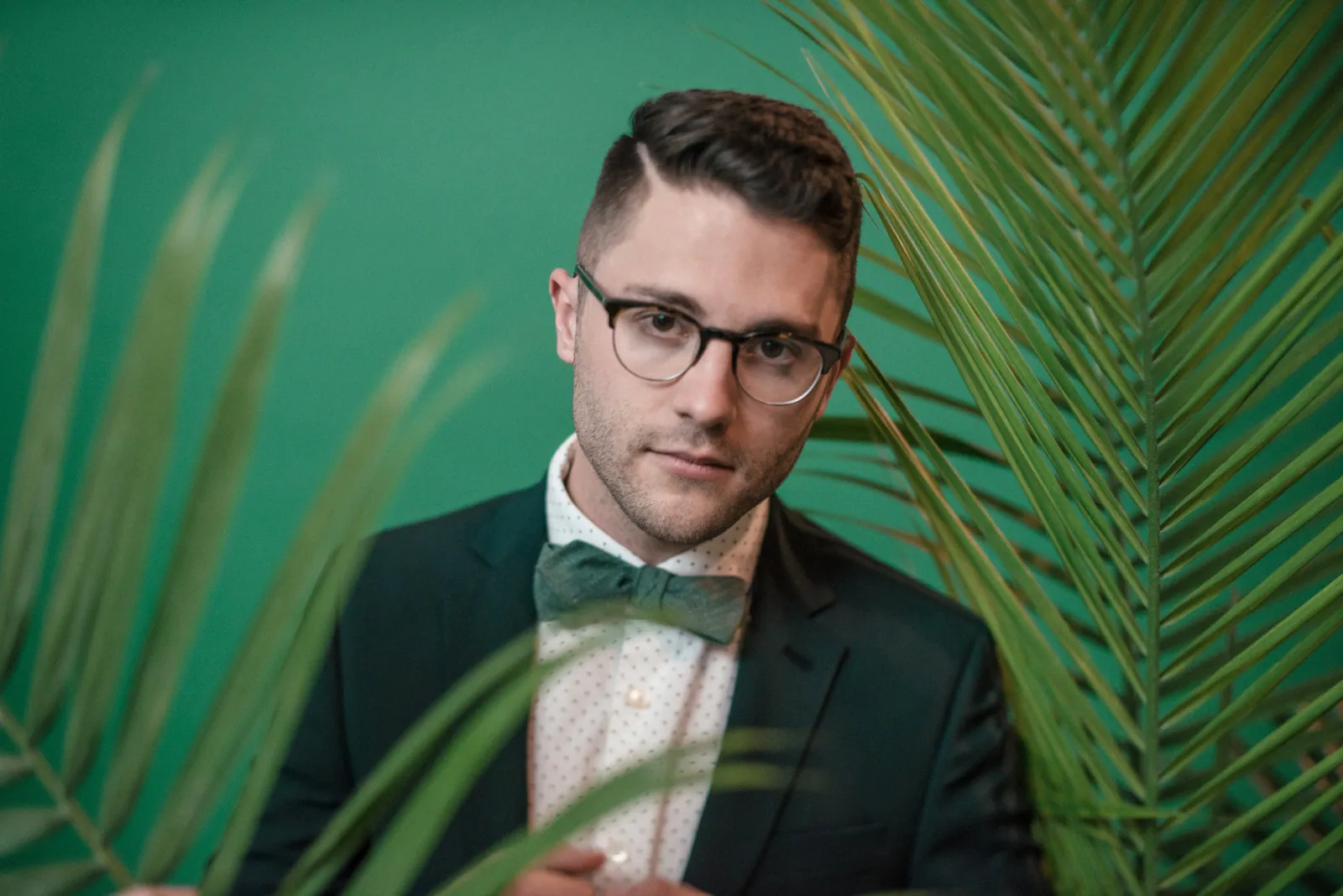 A man in dark-rimmed glasses, a green bow tie, and a dark blazer poses behind palm fronds against a green backdrop.
