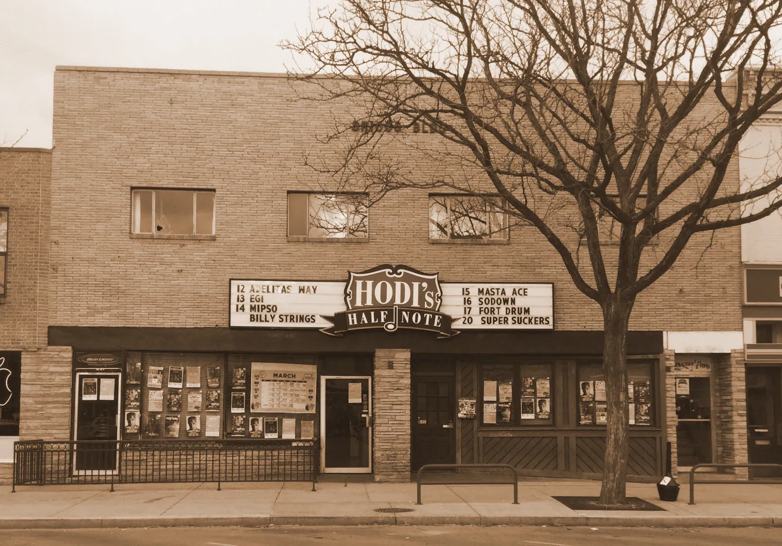 Sepia photo of Hodi's Half Note's brick storefront, with a marquee listing bands and a bare tree out front.