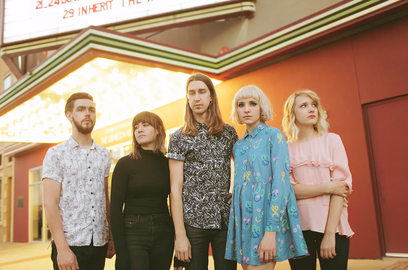 Five people stand in a row in front of a lit theater marquee, dressed in casual patterned and pastel outfits.