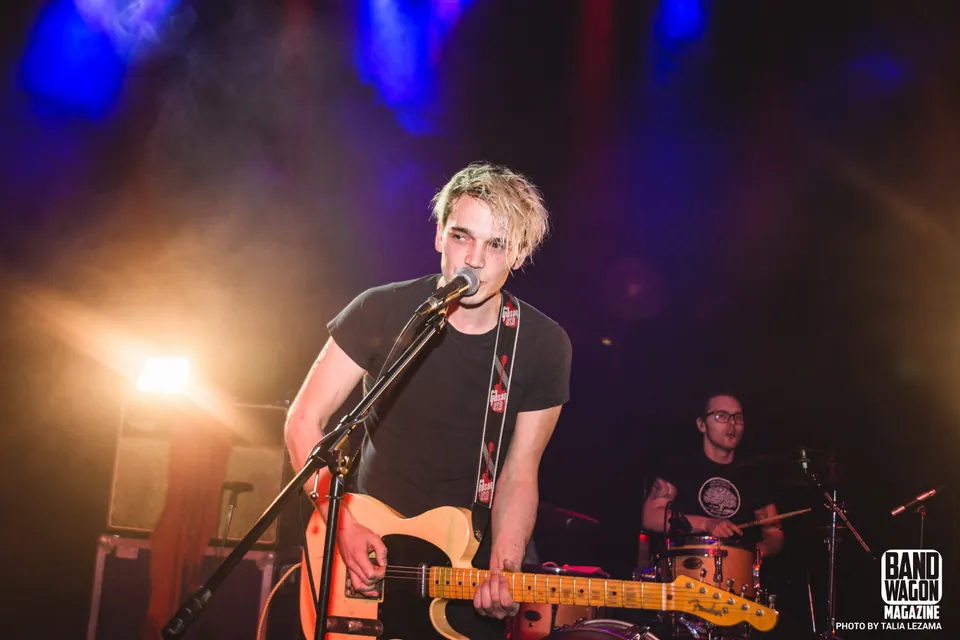 Blond singer in a black tee plays guitar at a mic as a bespectacled drummer performs under blue stage lights.