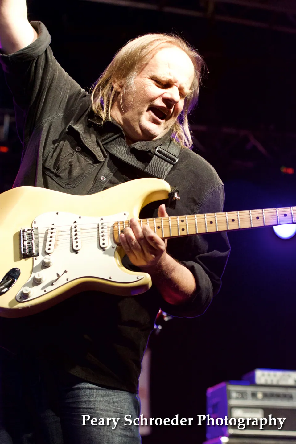 A long-haired guitarist in a black shirt plays a cream electric guitar with one arm raised on a dark stage.